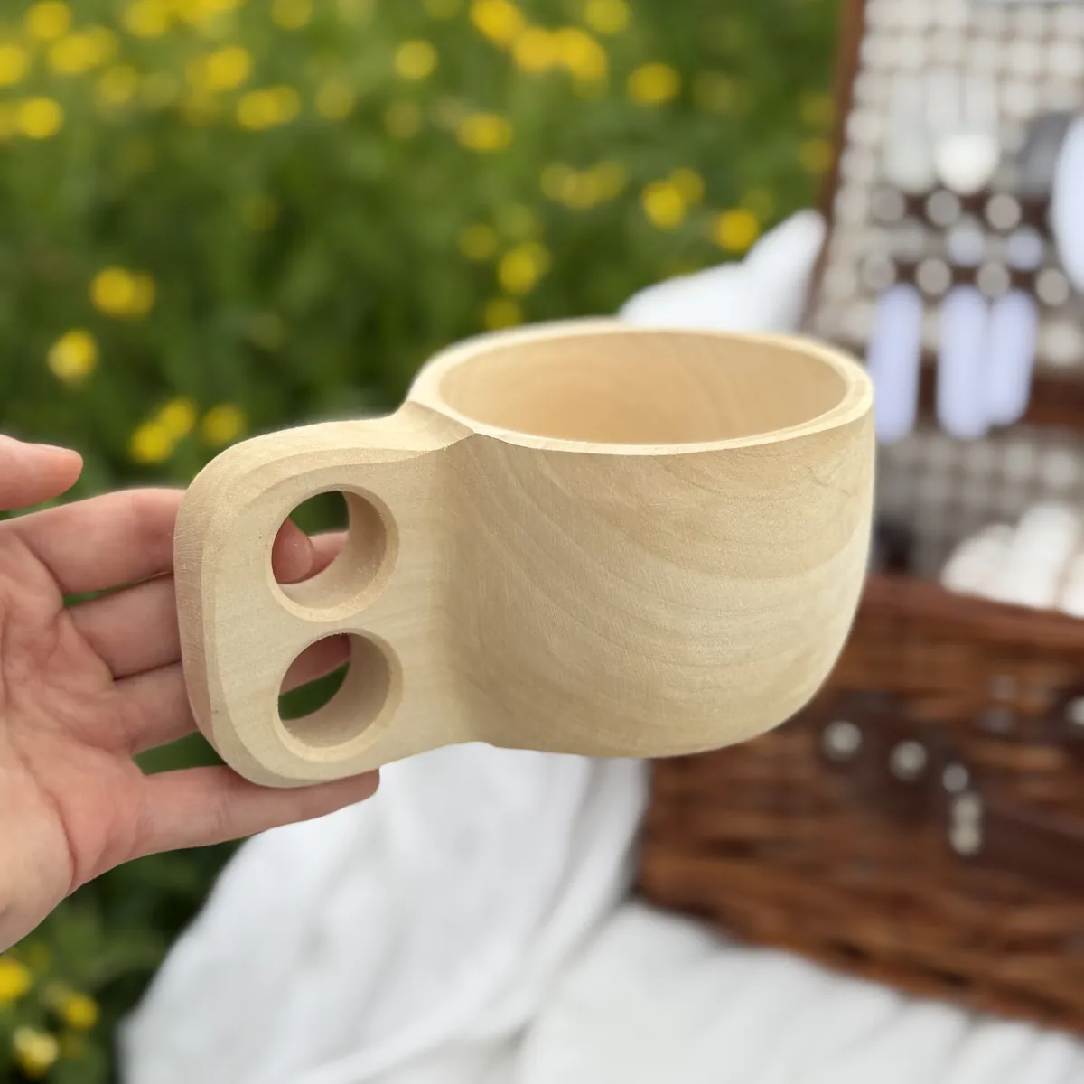 Kuksa z drewna bukowego trzymana w dłoni na tle kosza piknikowego