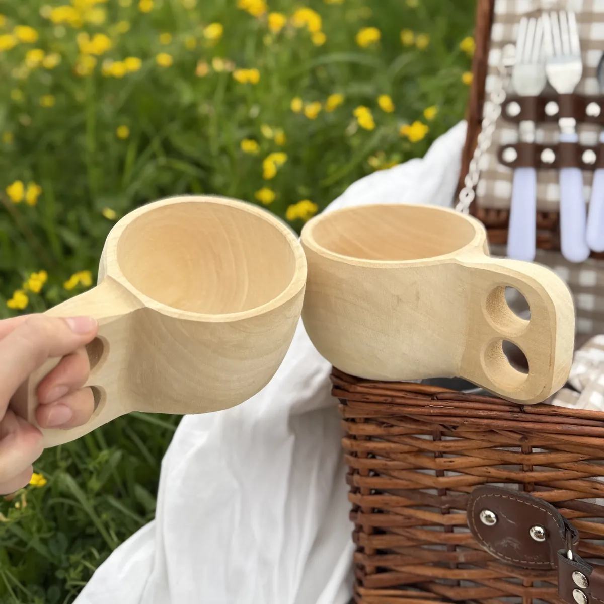Dwa bukowe kubki Kuksa z uchwytami na palce przy koszu piknikowym na łące